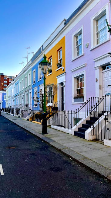 Image showing a row of colorful terraced houses on Fulham Palace Road, painted in pastel shades including light blue, yellow, pink, and lavender, with white window frames and black iron railings along the front steps. A traditional green street lamp is situated on the sidewalk near the houses, which have small front gardens enclosed by iron fencing. The pavement is made of concrete slabs, and the street appears quiet with no visible vehicles or pedestrians. This location highlights a typical residential area in West Kensington, suitable for home relocation or moving services provided by Man with Van West Kensington. The bright lighting indicates daylight, with a clear blue sky overhead, and the scene captures the external environment involved in packing, loading, and transporting furniture and household items during a same-day removal or house move.