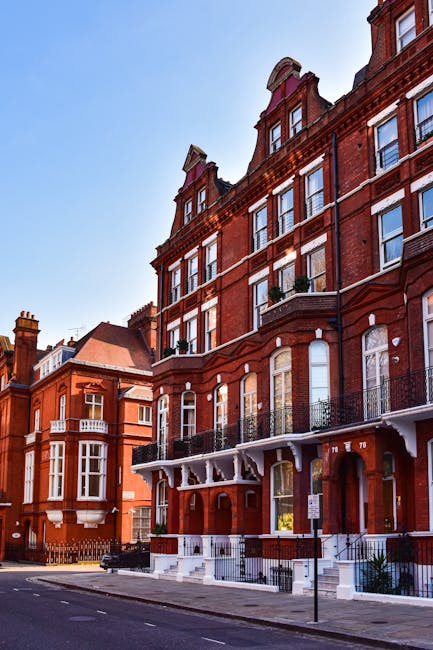 A line of red-brick Victorian-style residential buildings on Fulham Palace Road, with large sash windows, decorative iron balconies, and white-painted lower façades. The buildings are situated along a paved street with a few parked vehicles visible on the pavement. Clear blue sky above indicates a bright day. In the foreground, a man with Van West Kensington is preparing for a home relocation, with a large moving truck parked nearby. Visible in the image are cardboard boxes, wrapped furniture, and moving blankets on the pavement, suggesting an active furniture transport and packing process as part of the same-day removals service. The scene captures the typical urban environment of West Kensington during a relocation, with the loading process underway to facilitate efficient house removals from the property on Fulham Palace Road.
