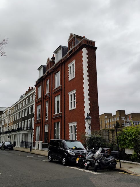Image showing a row of colorful terraced houses on Fulham Palace Road, painted in pastel shades including light blue, yellow, pink, and lavender, with white window frames and black iron railings along the front steps. A traditional green street lamp is situated on the sidewalk near the houses, which have small front gardens enclosed by iron fencing. The pavement is made of concrete slabs, and the street appears quiet with no visible vehicles or pedestrians. This location highlights a typical residential area in West Kensington, suitable for home relocation or moving services provided by Man with Van West Kensington. The bright lighting indicates daylight, with a clear blue sky overhead, and the scene captures the external environment involved in packing, loading, and transporting furniture and household items during a same-day removal or house move.