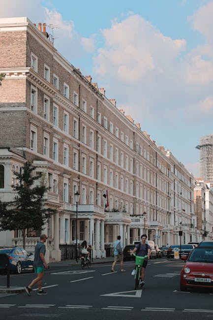 Image showing a spacious residential street lined with multi-storey Victorian-style terraced houses featuring detailed stonework and large sash windows, under a bright blue sky with some clouds. In the foreground, a man wearing casual clothing is riding a bicycle on the pavement, while another person walking nearby is pushing a cart or trolley, possibly used for moving purposes. Several parked cars are visible along the curb, including a red hatchback and a blue vehicle. The street appears busy with pedestrians and cyclists, and some construction scaffolding is visible in the background, indicating ongoing development or renovation work. The scene captures an urban environment suitable for house removal activities, with potential for loading and unloading furniture and boxes from nearby properties. Natural daylight enhances the clarity of the environment, supporting the context of home relocation and furniture transport. The image aligns with house removals services as offered by Man with Van West Kensington, emphasizing efficient street access for moving logistics under allowed permit rules for W14, WEST KENSINGTON.