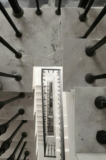 An overhead view of a narrow staircase inside a building showing a modern, minimalist design with light grey concrete steps and black metal balustrades. The staircase is viewed from the top looking downward, revealing multiple flights connected by small landings. The concrete steps are supported by black metal rods, and the surrounding area appears clean and well-maintained. The image captures the vertical extent of the staircase, emphasizing its compact, tight spiral structure, which may present challenges during home relocation or furniture transport. This type of staircase is often encountered in residential properties undergoing packing and moving procedures, where careful planning is needed for lifting and loading objects safely. Occasionally, moving services like those offered by Man with Van West Kensington may include navigating such narrow stairs as part of their expertise in house removals and logistical planning for efficient furniture transport within confined spaces.