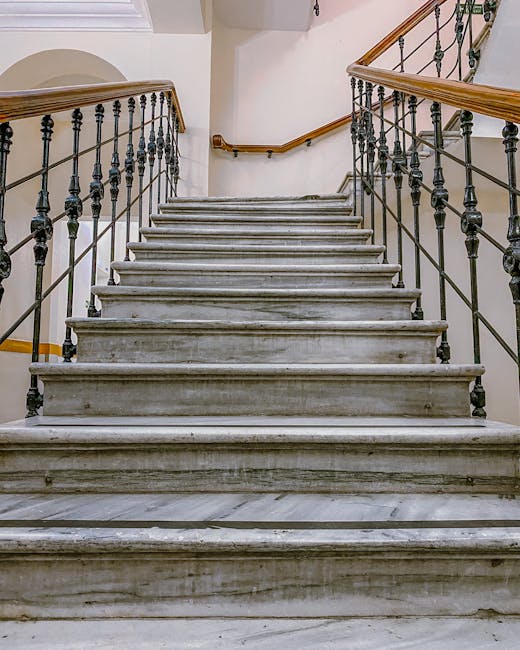 An overhead view of a narrow staircase inside a building showing a modern, minimalist design with light grey concrete steps and black metal balustrades. The staircase is viewed from the top looking downward, revealing multiple flights connected by small landings. The concrete steps are supported by black metal rods, and the surrounding area appears clean and well-maintained. The image captures the vertical extent of the staircase, emphasizing its compact, tight spiral structure, which may present challenges during home relocation or furniture transport. This type of staircase is often encountered in residential properties undergoing packing and moving procedures, where careful planning is needed for lifting and loading objects safely. Occasionally, moving services like those offered by Man with Van West Kensington may include navigating such narrow stairs as part of their expertise in house removals and logistical planning for efficient furniture transport within confined spaces.