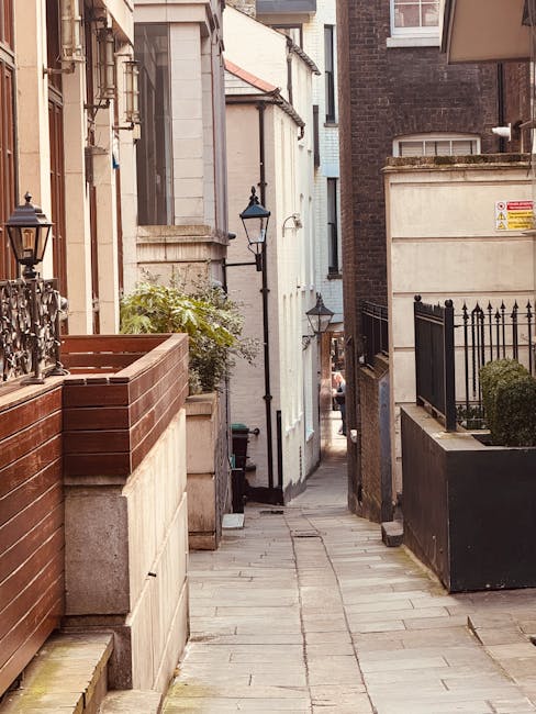 A narrow, urban residential alleyway with a paved stone pathway, flanked by low brick and concrete walls topped with metal railings on both sides. On the left, there is a wooden planter box and a decorative lantern attached to the wall. On the right, a large black metal gate is visible, along with a small bush in a container. The alley is lined with traditional-style street lamps attached to the buildings, leading towards a view of additional narrow alleyways and building facades in the distance. The scene is well-lit with natural daylight, highlighting the textures of the brick, concrete, and paving stones. This setting reflects the challenges of home relocation and furniture transport through confined spaces, as handled by professional removals specialists such as Man with Van West Kensington, especially when dealing with properties on North End Road with narrow access.