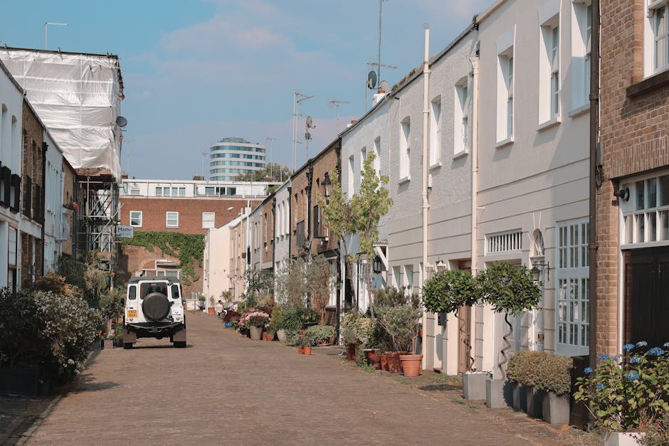 A narrow residential street in West Kensington with cobbled pavement and row houses on both sides, some with brick facades and others with white exterior finishes. Several potted plants and small trees are positioned along the front of the houses, adding greenery to the scene. A white van belonging to Man with Van West Kensington is parked on the street, suggesting a home relocation or furniture transport process. The van's side door is closed, and the street appears quiet with no moving activity visible at this moment. In the background, there is a modern, multi-storey building with curved architecture, contrasting with the traditional houses in the foreground. The scene is lit with natural daylight, with a partly cloudy sky overhead, indicating daytime conditions suitable for packing and loading furniture for a move in a property with narrow access. This setting reflects the logistical considerations of house removals in tight spaces, highlighting the importance of specialized transportation solutions for such environments.