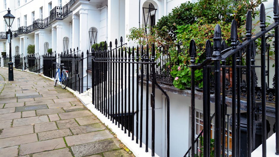 A row of white terraced houses with black decorative wrought iron railings in front, lining a paved sidewalk made of rectangular stone slabs. A blue bicycle leans against the railings near the entrance of one property. The building facades feature large windows with black metal frames and some have small balconies. Green leafy plants and pink flowers are visible on the right side, growing along the railings and climbing the building walls. The image is well-lit with natural daylight, capturing an urban neighbourhood scene in West Kensington that is relevant to house removals and moving services, such as loading or transportation preparations. The setting suggests a residential street where furniture transport and home relocation activities might take place, with the environment indicating a clean, orderly exterior suitable for professional removals by Man with Van West Kensington.