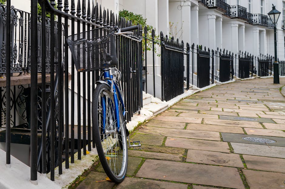 A row of white terraced houses with black decorative wrought iron railings in front, lining a paved sidewalk made of rectangular stone slabs. A blue bicycle leans against the railings near the entrance of one property. The building facades feature large windows with black metal frames and some have small balconies. Green leafy plants and pink flowers are visible on the right side, growing along the railings and climbing the building walls. The image is well-lit with natural daylight, capturing an urban neighbourhood scene in West Kensington that is relevant to house removals and moving services, such as loading or transportation preparations. The setting suggests a residential street where furniture transport and home relocation activities might take place, with the environment indicating a clean, orderly exterior suitable for professional removals by Man with Van West Kensington.